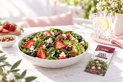 A fresh green salad in a white bowl on a sunlit outdoor table, with a packet of Pocket Flavors Very Berry flavor seasoning mix placed beside it. The salad includes spinach, strawberries, blueberries, pecans, and avocado.