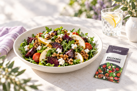 A fresh salad in a white bowl on a sunlit table, with a packet of Pocket Flavors Balsamic Seasoning placed beside it. The salad includes arugula, sliced beets, pears, walnuts, and crumbled cheese, with a glass of lemonade and greenery in the background.