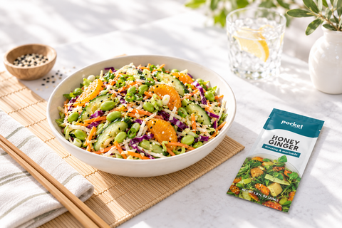 A bowl of fresh salad with cucumber, shredded cabbage, edamame, carrots, orange slices, and dressing sits on a bamboo mat. A small packet labeled pocket Honey Ginger salad dressing is placed beside the bowl on a white table, with a glass of lemonade and a small bowl of peppercorns in the background.