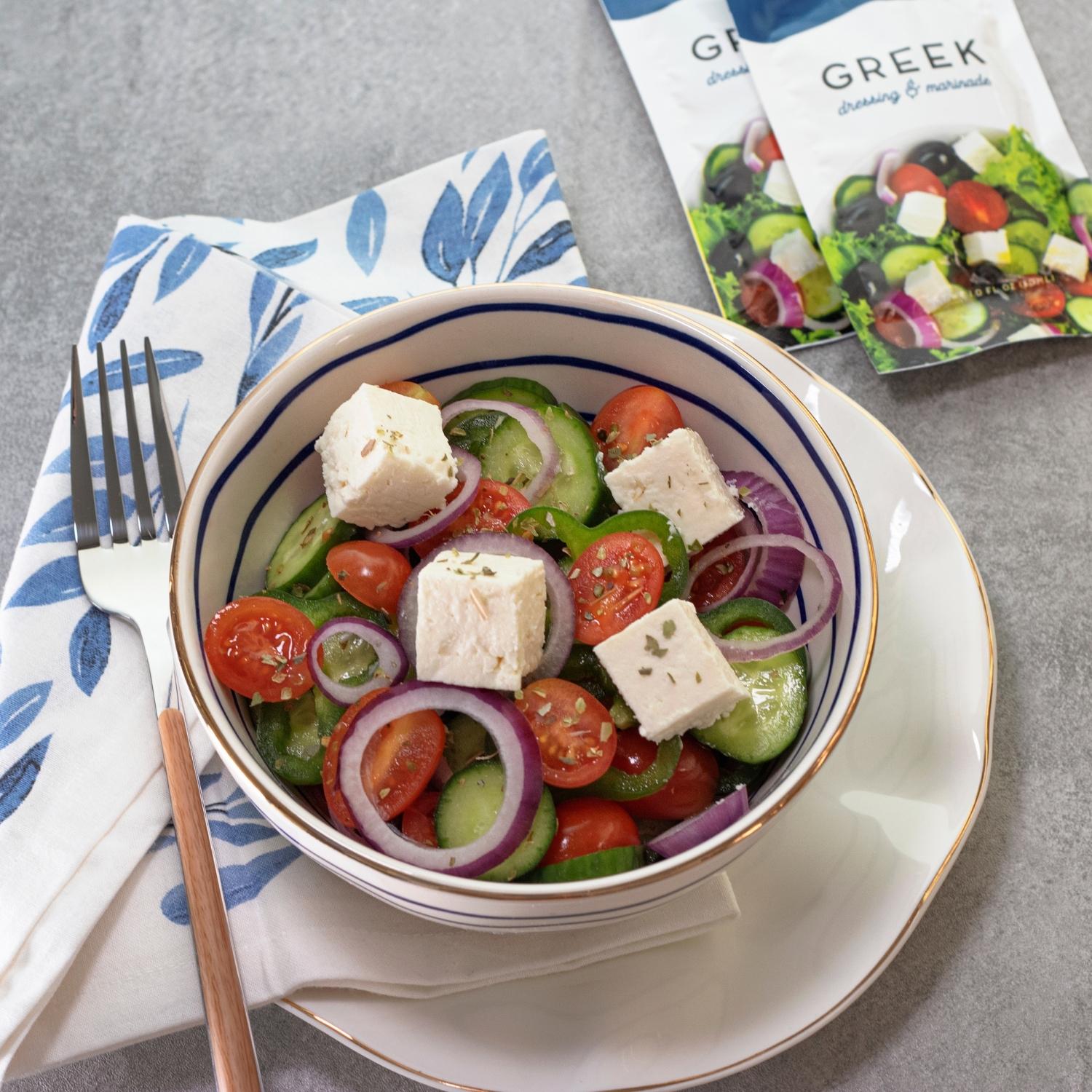 Single serve greek salad dressing packet next to greek salad bowl with feta, tomatoes, red onion, and cucumber.